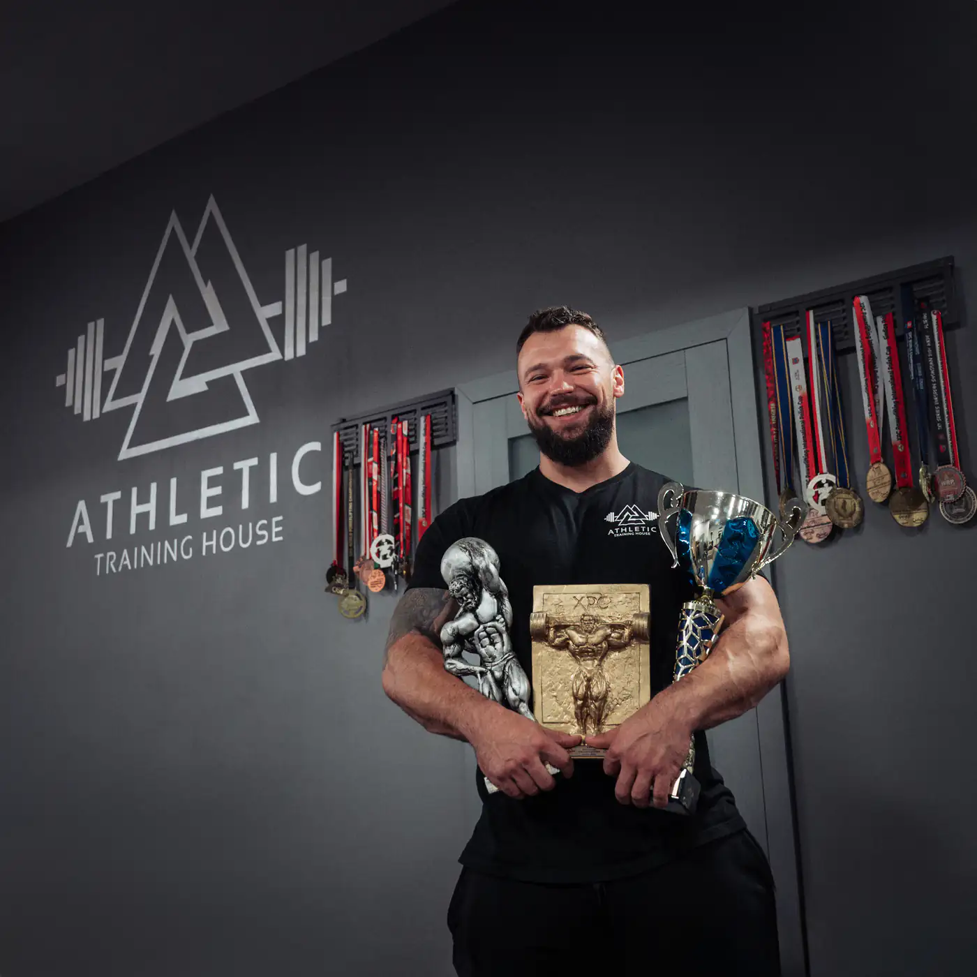 Mariusz Marszalik z pucharami i statuetką XPC przed logo Athletic Training House i ścianą z medalami